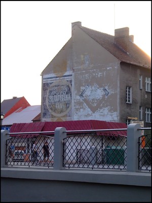 PRAKTYCZNA PANI. Autor: Łukasz Wojciak. Zdjęcie przedstawia starą elewację budynku widzianą w tle odnowionego mostu na ul. Piłsudskiego